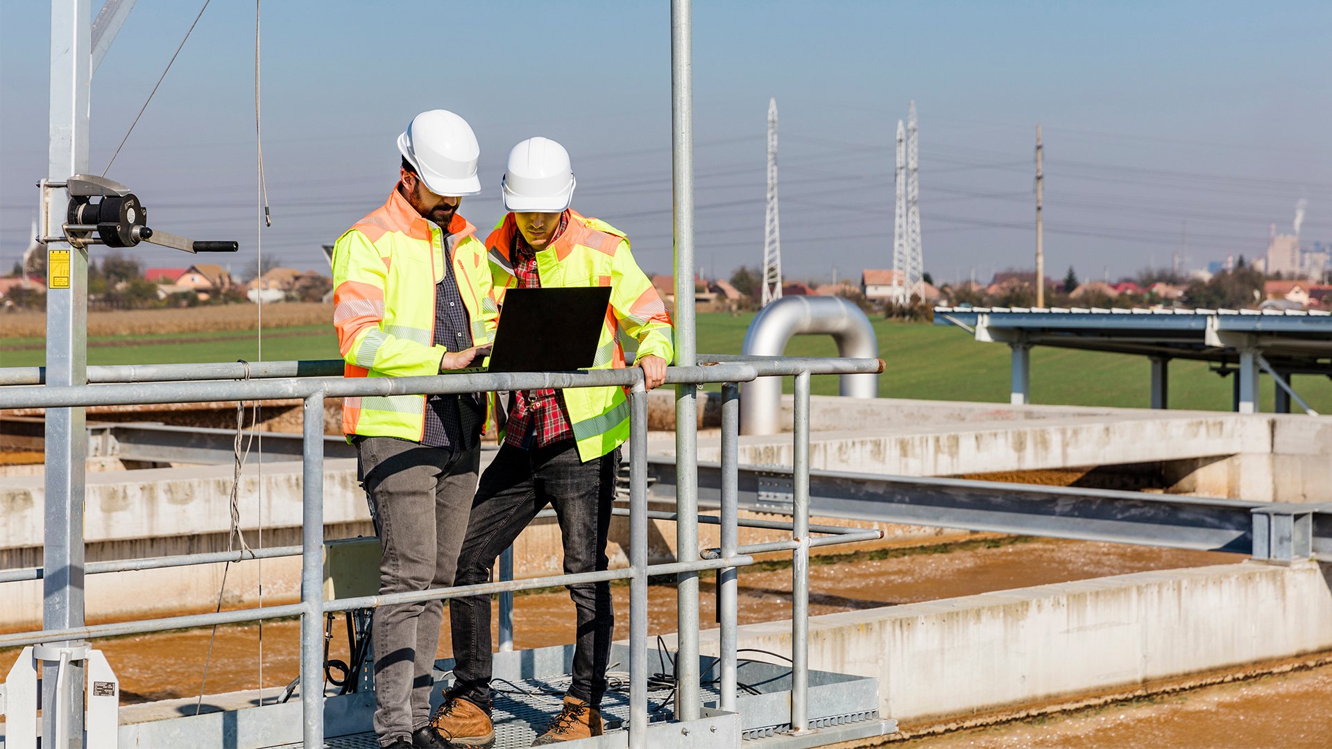 Two men wearing white hard hats and yellow and orange safety jackets standing outside at a water plant entering information into a laptop.
