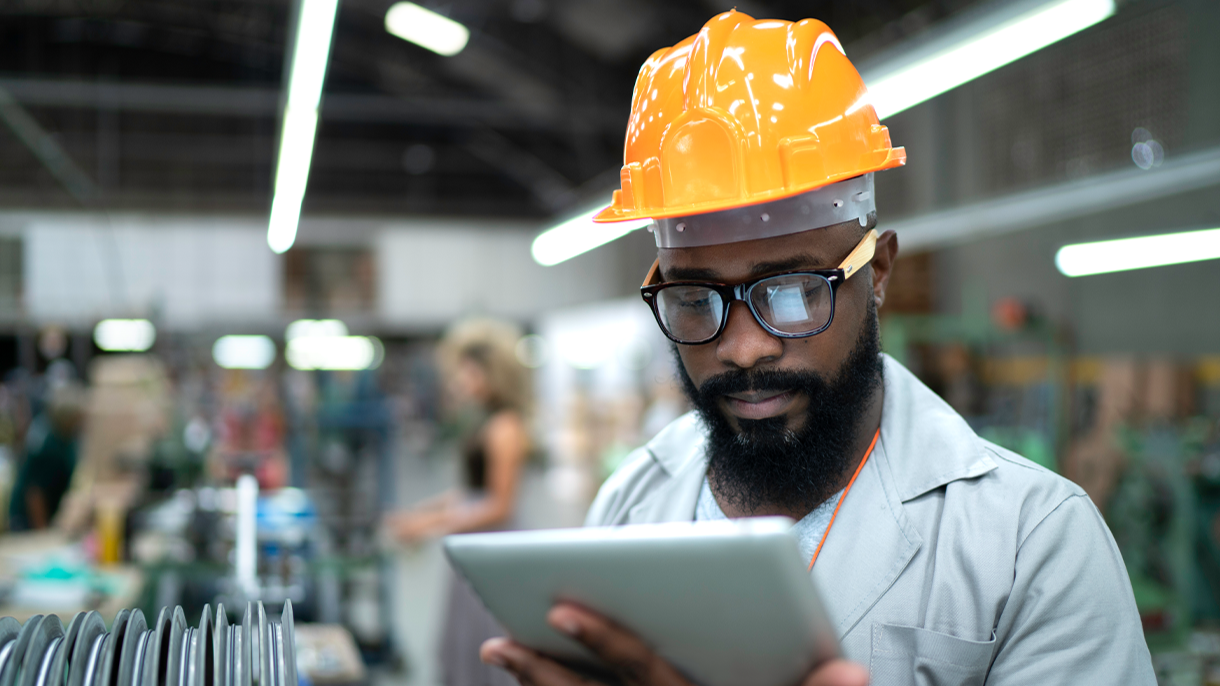 A man in a hard hat and glasses is focused on using a tablet at a construction site
