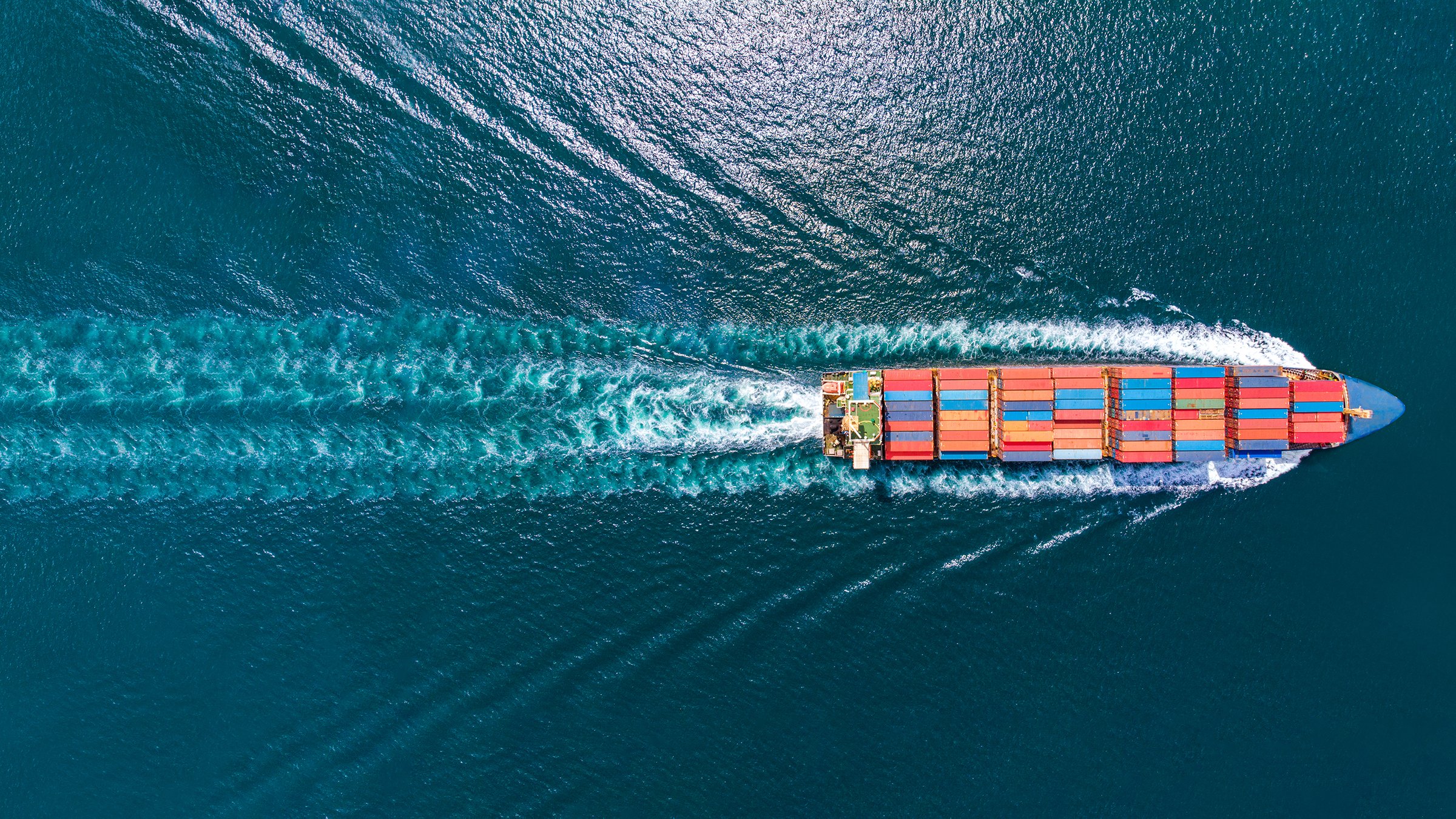 Aerial top view of cargo ship carrying containers freight in the ocean with a wake behind it 