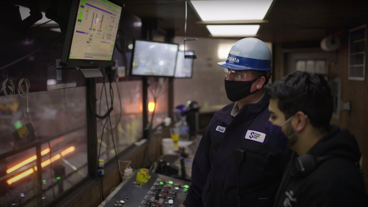 Two steel workers wearing masks and one in a hard hat look at a screen to evaluate the status of their operations in a control room.