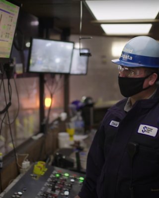 Two steel workers wearing masks and one in a hard hat look at a screen to evaluate the status of their operations in a control room.