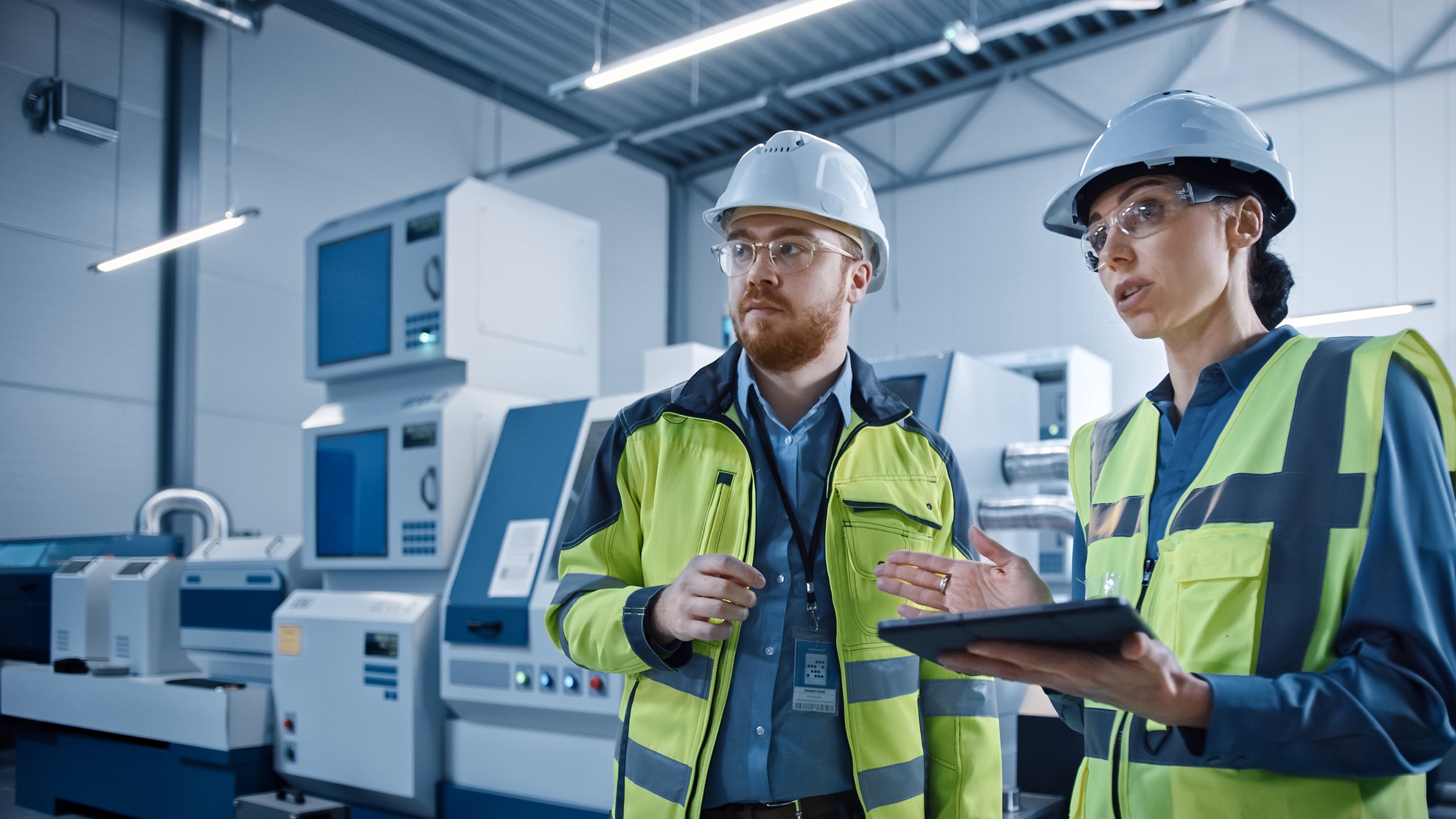 Une cheffe de projet et un ingénieur debout, portant des gilets de sécurité et des casques de protection, travaillent dans un atelier, discutent, utilisent une tablette numérique et surveillent une chaîne de montage de machines CNC, dans une usine moderne : Une cheffe de projet et un ingénieur debout, portant des gilets de sécurité et des casques de protection, travaillent dans un atelier, discutent, utilisent une tablette numérique et surveillent une chaîne de montage de machines CNC