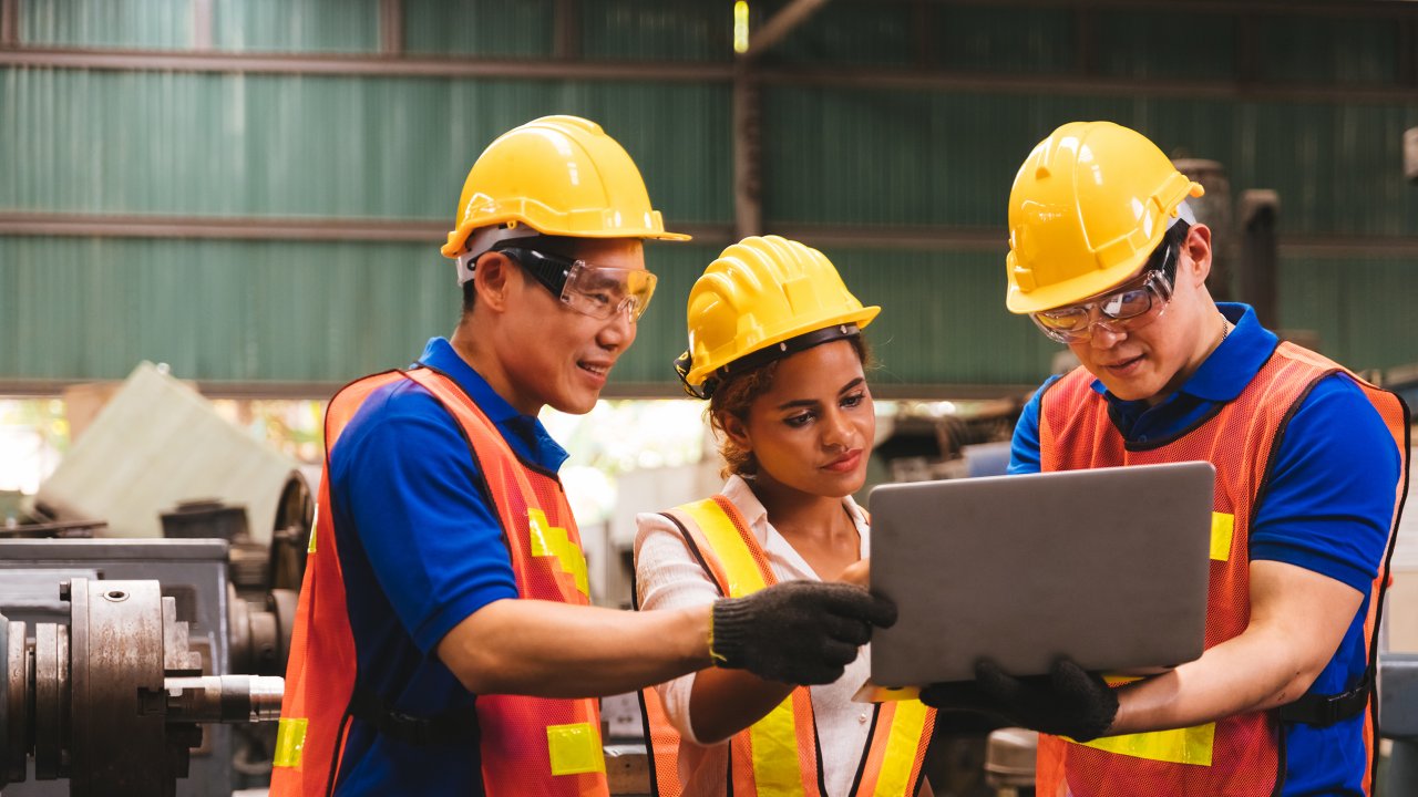Un ingeniero técnico asiático y una ingeniera de piel morena con uniformes y cascos de seguridad en una inspección, controlando el proceso de producción con una computadora portátil en una planta.
