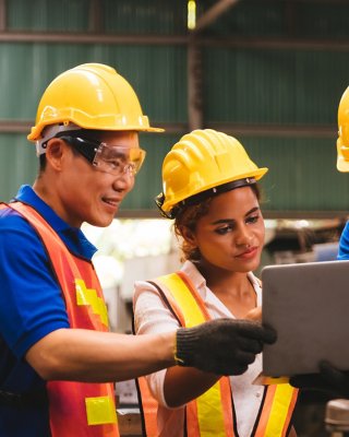 ユニフォームと安全ヘルメットを着用したアジア系の技術者と黒い肌の女性エンジニアが、工場のステーションでラップトップコンピュータを使って生産工程を検査・確認している。
