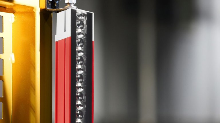 Una vista ravvicinata di un terzo superiore di una barriera fotoelettrica di sicurezza GuardShield 450L rossa, con terminazione grigia, montata lateralmente su una macchina metallica gialla con uno sfondo sfocato in grigio. Il lato della lente ottica è rivolto a destra.
