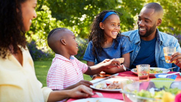 Quatro pessoas de uma família sentadas juntas à uma mesa de piquenique, comendo e rindo