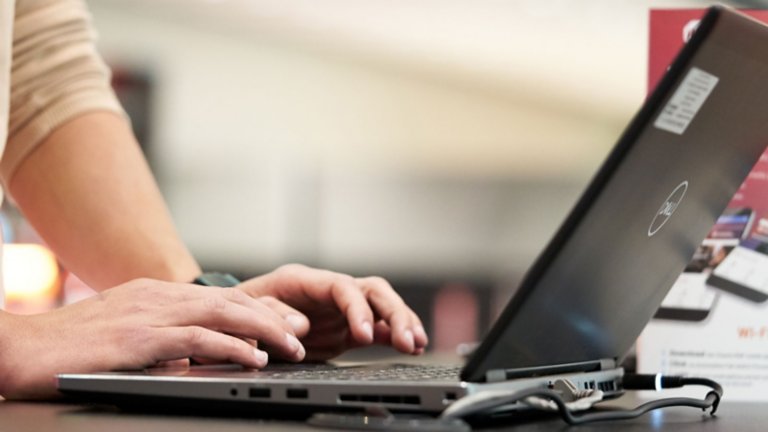 Zbliżenie na ręce młodej osoby piszącej na klawiaturze czarnego laptopa w środowisku biurowym i poznającej możliwości oprogramowania Connected Components Workbench w chmurowej platformie szkoleniowo-prezentacyjnej firmy Rockwell Automation. Laptop jest włączony i podłączony do źródła zasilania. 