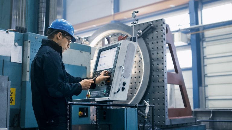 Funcionário usando um capacete azul digitando informações em um monitor que está conectado a uma máquina em uma fábrica