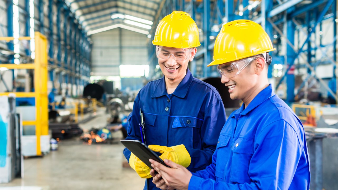 Two manufacturing workers in goggles and hard hats look at a tablet.