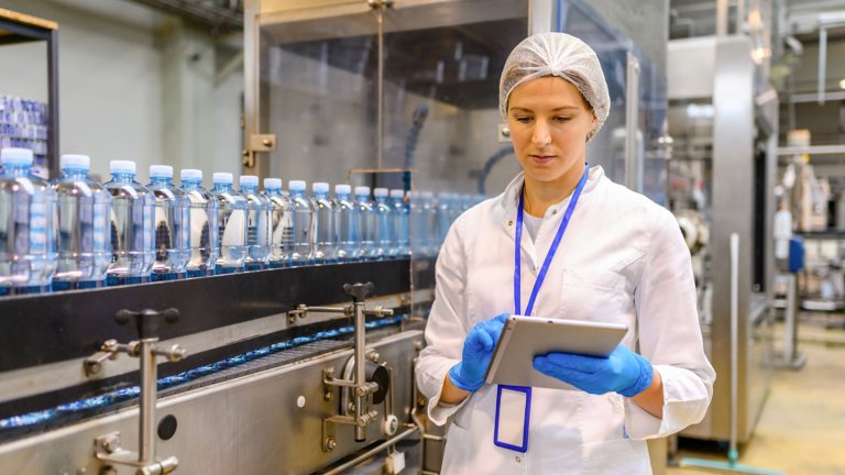 Female engineer analyzing the CPGSuite software on a tablet with a machine manufacturing bottles behind her