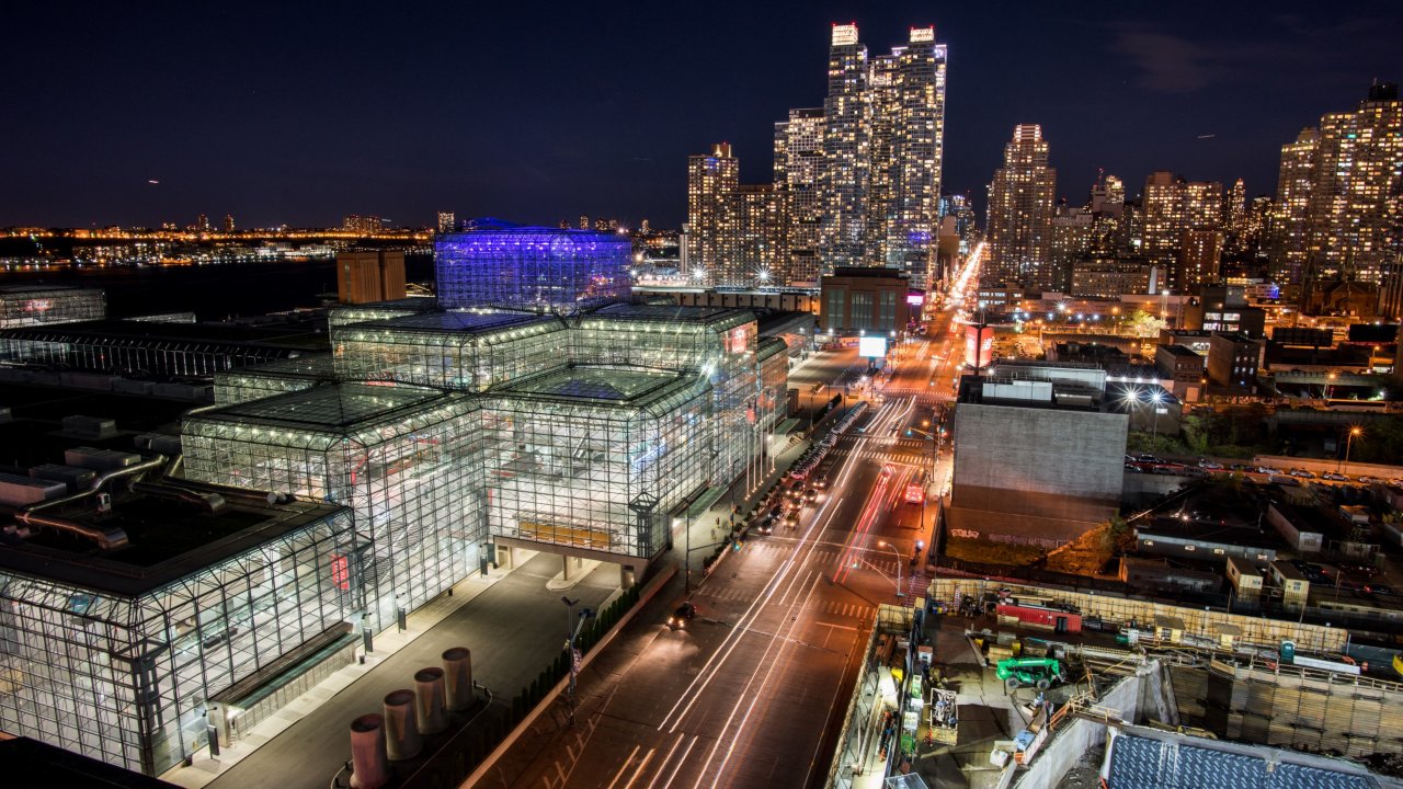An overhead perspective of the Javits Center illuminated at night, with the New York City skyline behind it.