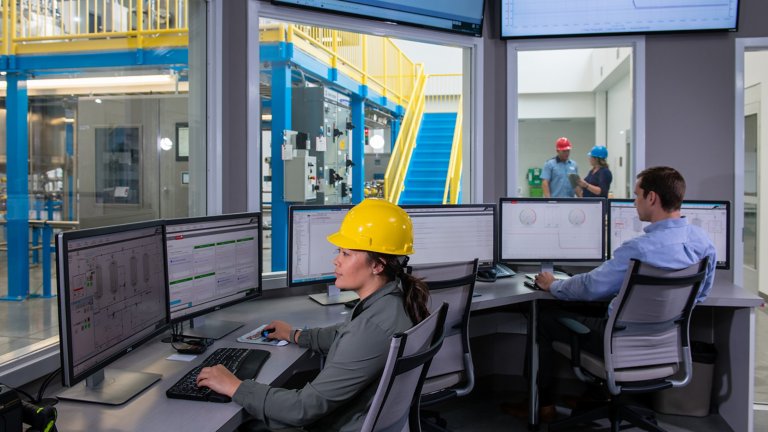 Um funcionário e uma funcionária usando capacetes amarelos, sentados a uma mesa de canto com vários monitores, em um escritório com janelas voltadas para a fábrica