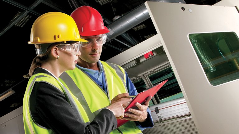 A woman wearing a yellow hard hat, safety glasses and a yellow reflective safety vest talking to a man wearing wearing a red hard hat, safety glasses and a yellow reflective safety vest.  Both are looking at papers on a red clip board while standing in front of industrial machinery.