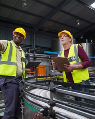 Two plant workers stand near manufacturing line sharing information on tablet and clipboard