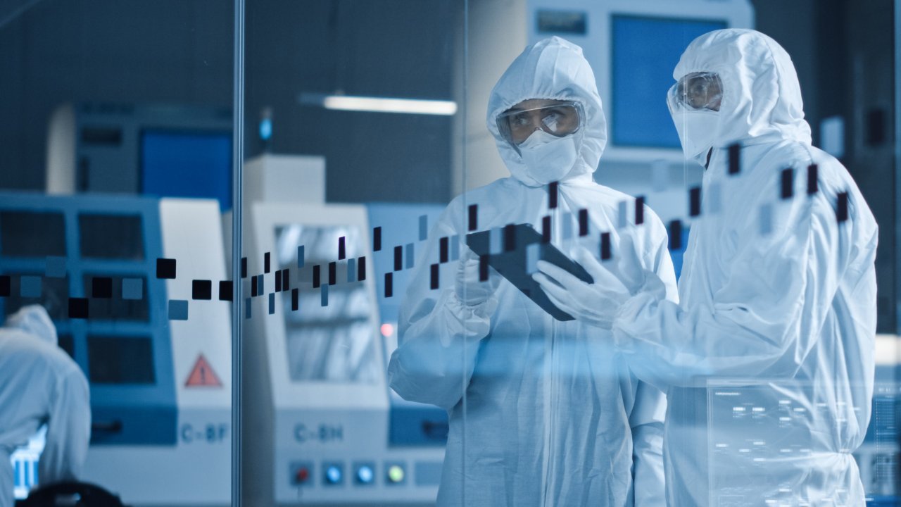 Dois profissionais de ciências da vida vestindo macacões e usando óculos de segurança revisam as análises em um tablet enquanto observam o processo de produção nas instalações da organização.