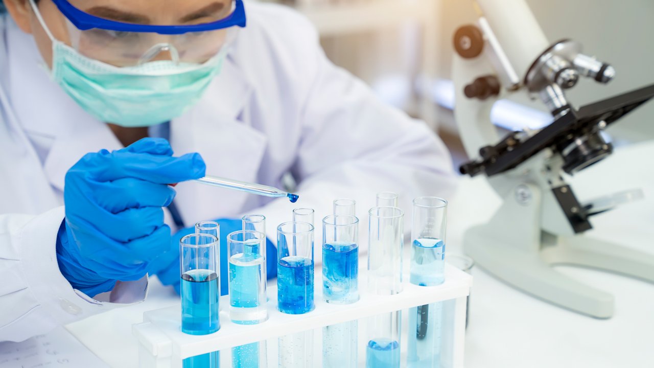 实验室内，穿着防护装备的男子在显微镜旁边的试管中滴入蓝色液体。