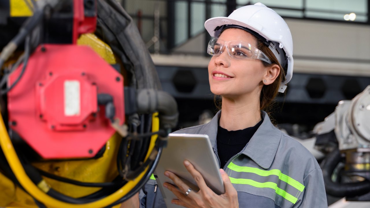 Female engineer in a white helmet in a production hall with tablet and robotic machine automation