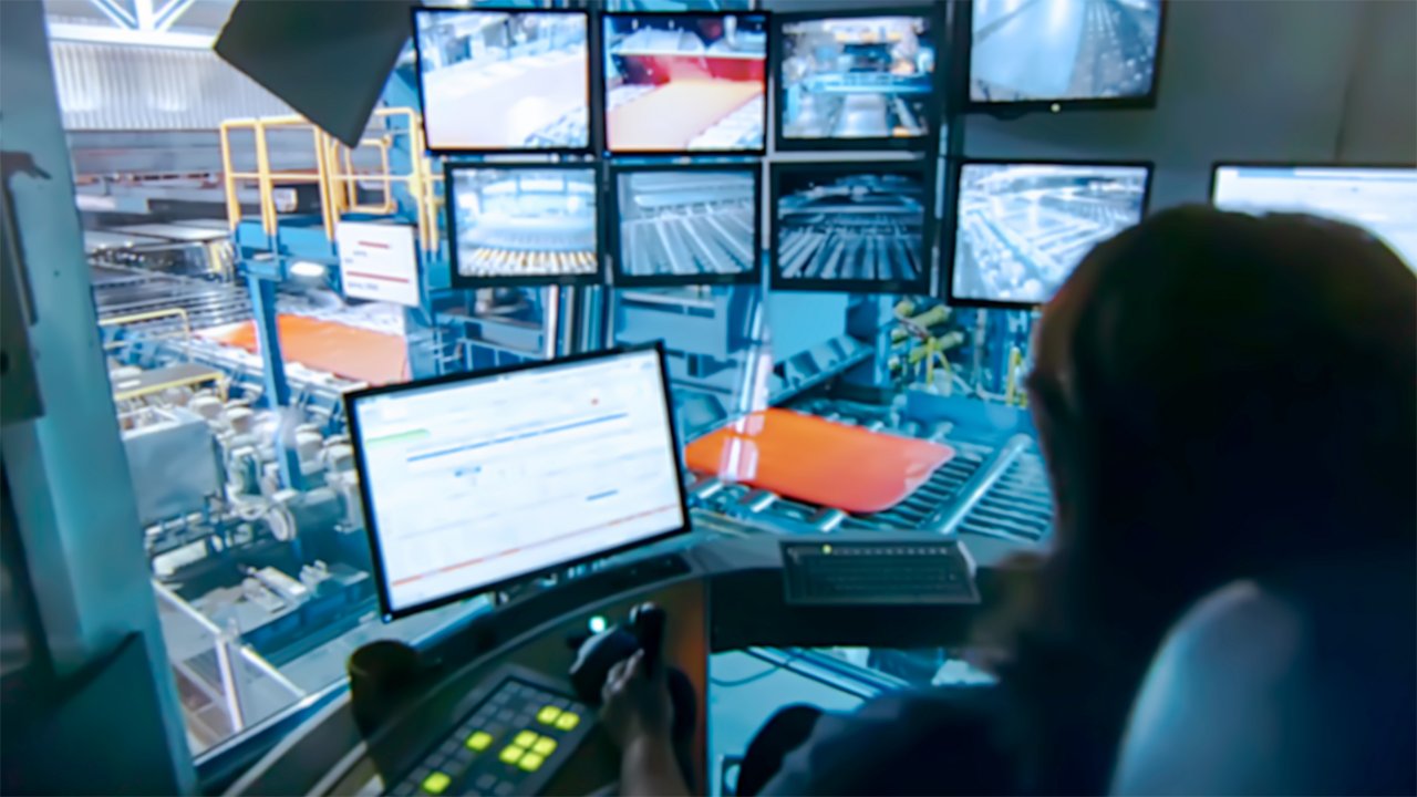 Empleada en una oficina observa una planta y ve información en su monitor con un número de monitores colgantes que muestran diferentes ubicaciones dentro de la planta a su derecha