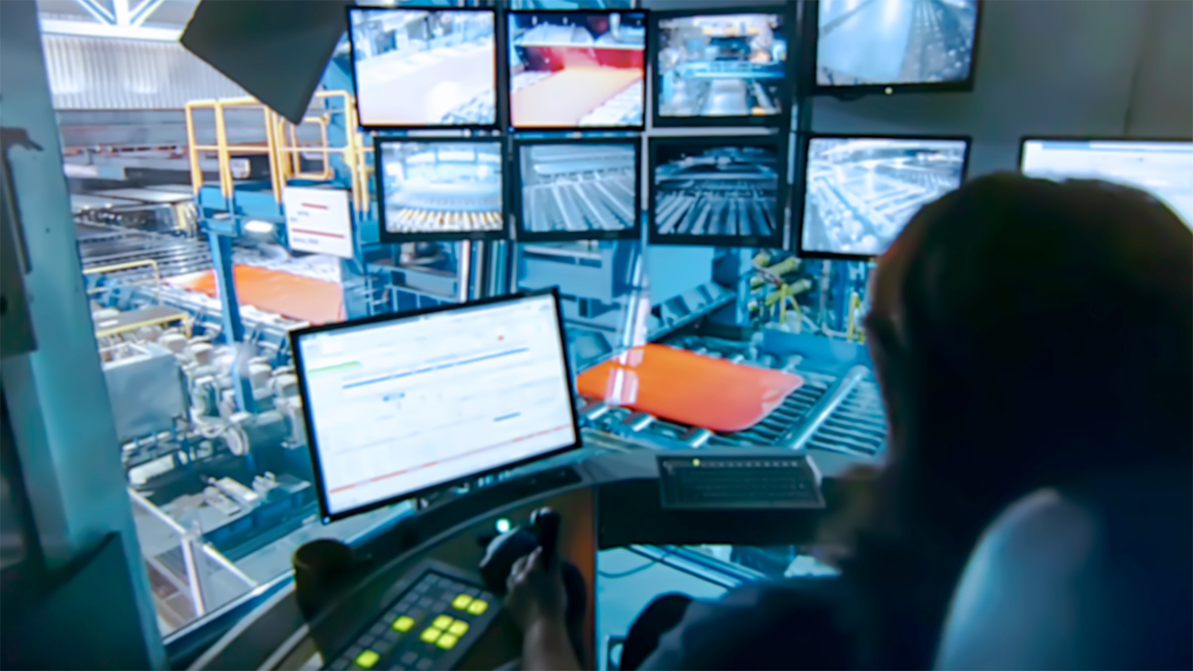 Female employee in a office that looks down at a plant floor viewing information on her monitor with a number of hanging monitors displaying locations within the plant to her right side