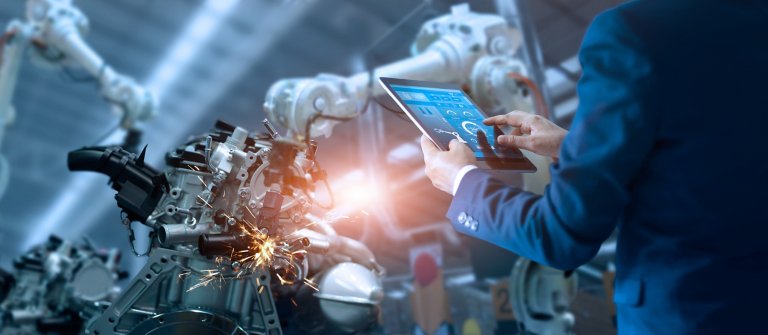 Pessoa em um terno azul usando um tablet com um software de fabricação na tela enquanto gerencia equipamentos industriais no chão de fábrica.