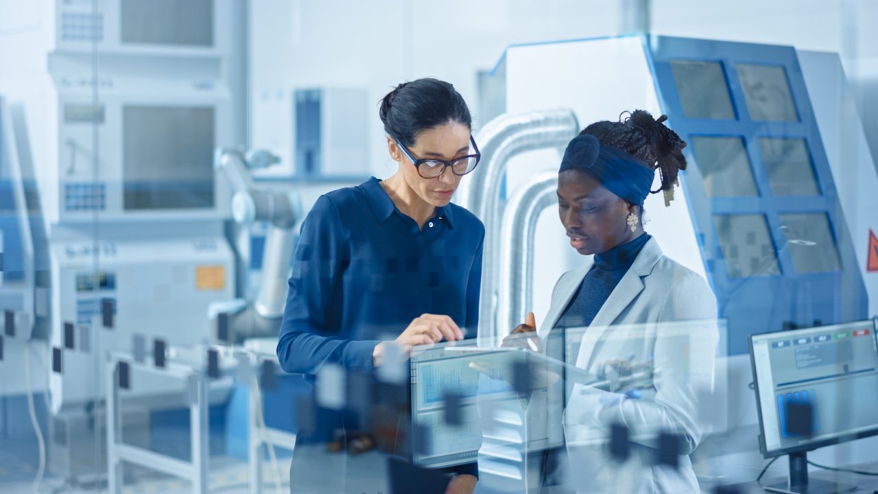 Two women dressed in business attire are in a manufacturing setting looking at a tablet.