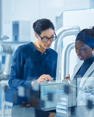 Two women dressed in business attire are in a manufacturing setting looking at a tablet.