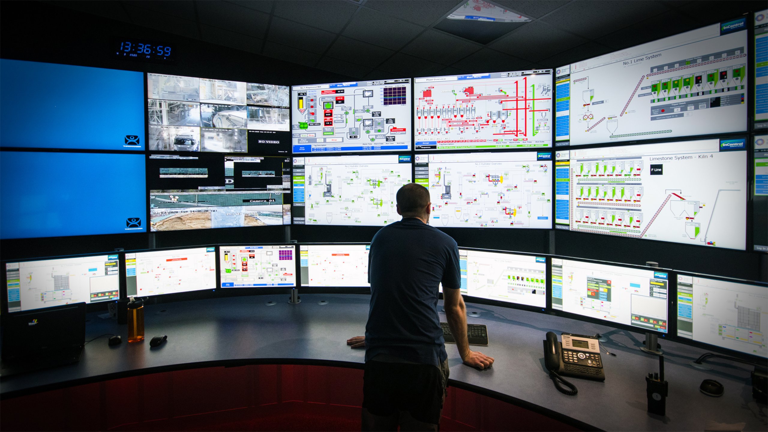 Ingeniero de pie frente a un gran escritorio curvado mirando varios monitores en una pared que incluyen una variedad de pantallas diferentes de una aplicación de software