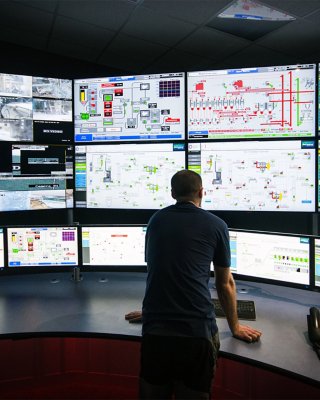 Ingenieur steht vor einem großen, geschwungenen Schreibtisch und blickt auf mehrere Monitore auf einer Wand, einschließlich einer Vielzahl verschiedener Bildschirme von einer Software-Anwendung