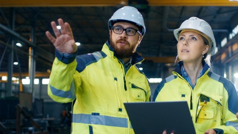 Man on left, woman on right in a manufacturing environment having a discussion while looking at a tablet. Both are wearing yellow safety coats and white hard hats.