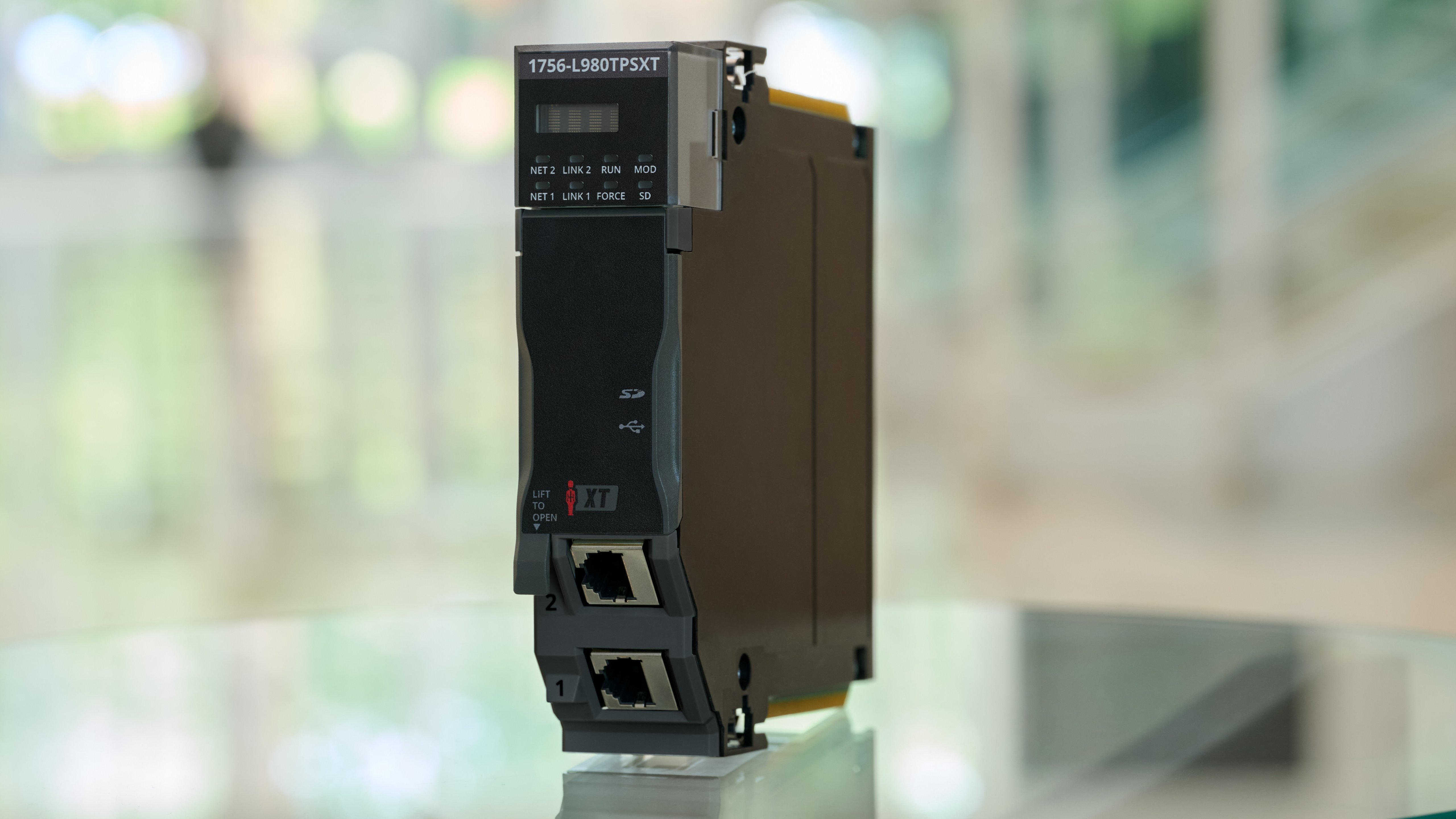 An industrial automation module labeled 1756-L98OTPSXT featuring dual Ethernet ports, multiple status indicators, and a front panel with “XT” branding. The module is photographed in a bright indoor setting with a blurred green and white background.