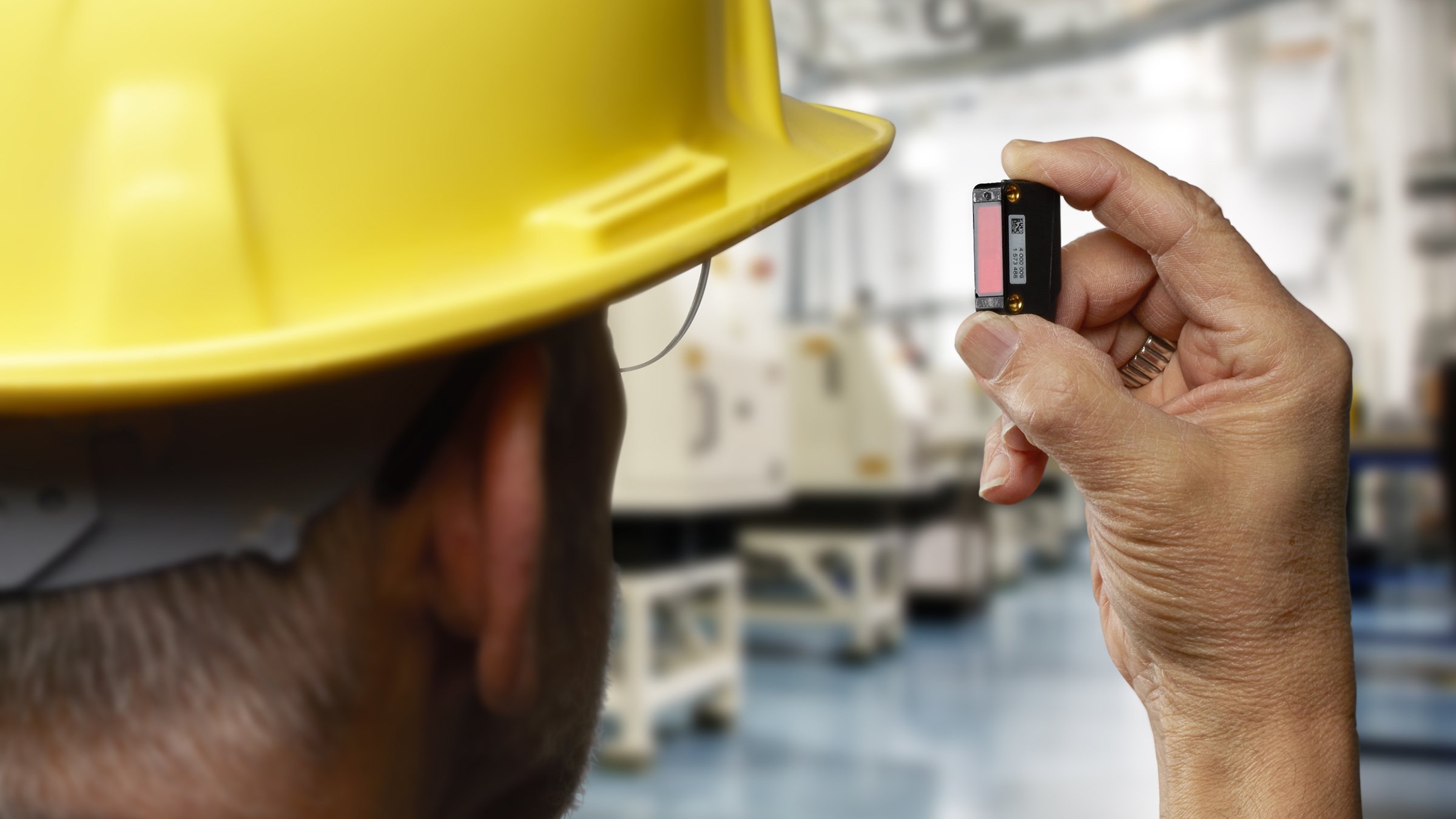 An over the shoulder view of a man in a yellow hard hat holding up a black Allen-Bradley 42JA VisiSight photoelectric sensor.