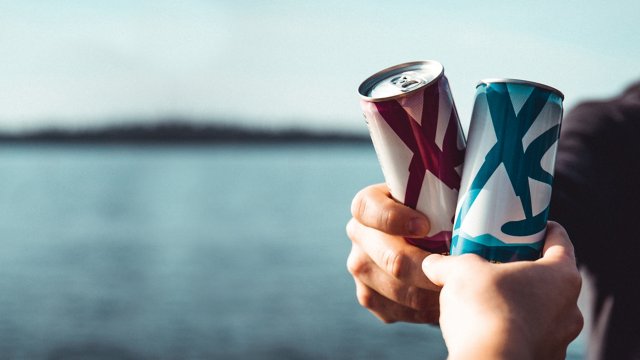 Dos manos con dos latas de bebida con logotipos delante de un paisaje acuático; la lata posterior es de color granate y la lata frontal es de color verde azulado.