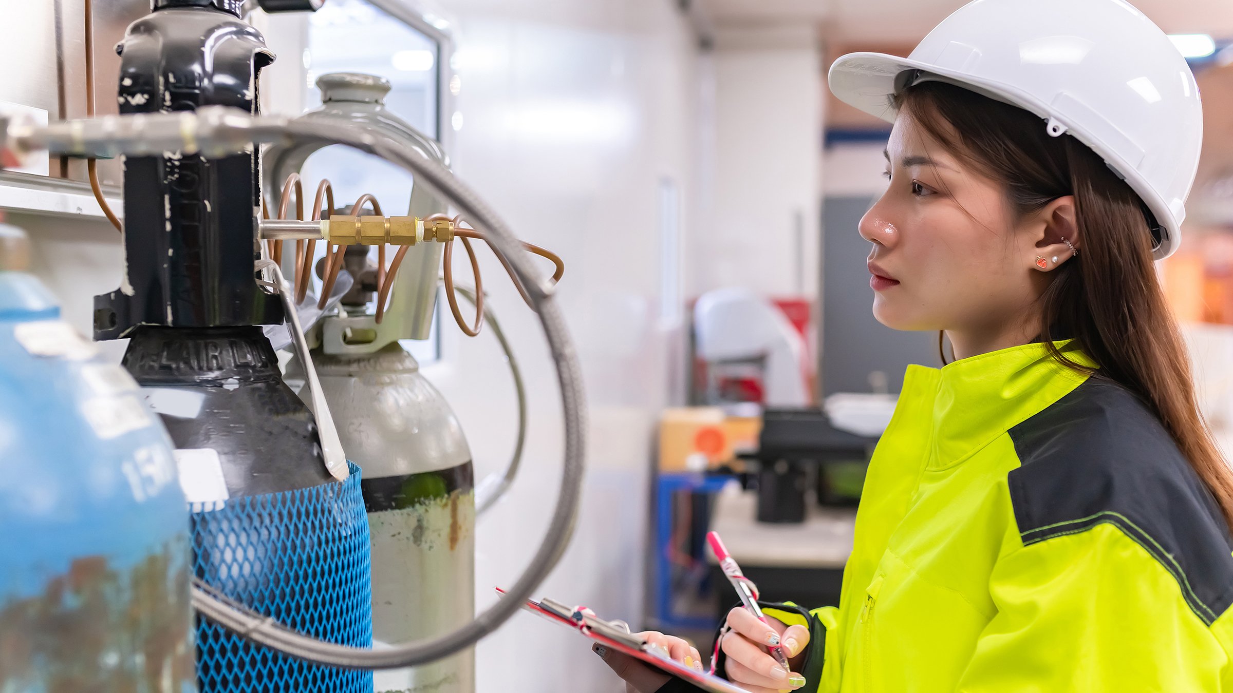 Female engineer in yellow safety gear with clipboard studying a piece of green hydrogen equipment.