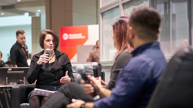 A woman is holding a microphone and talking to two people sitting on a couch at Automation Fair 2019