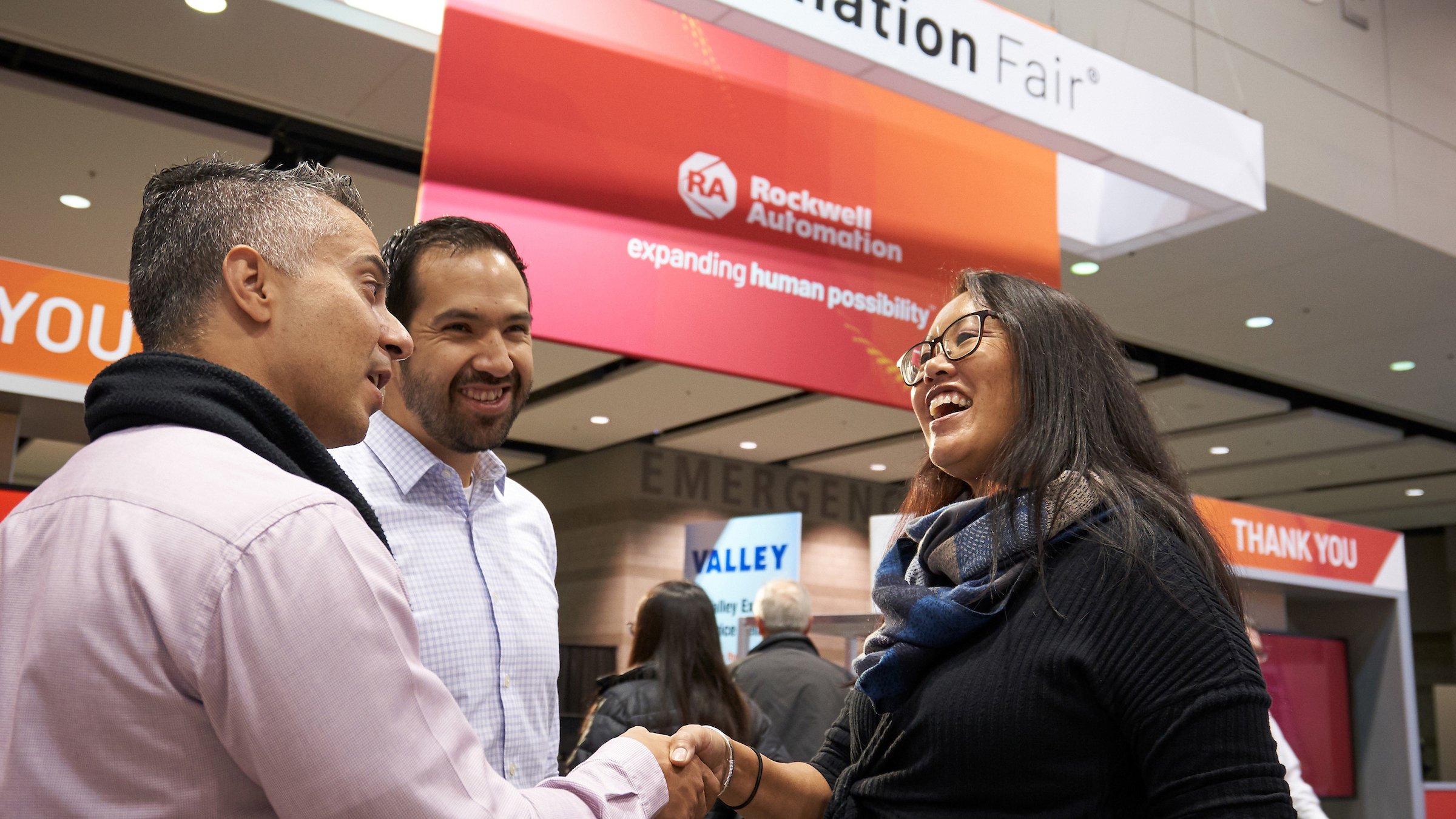 Two males and a female conversing and shaking hands with an Automation Fair banner in the background