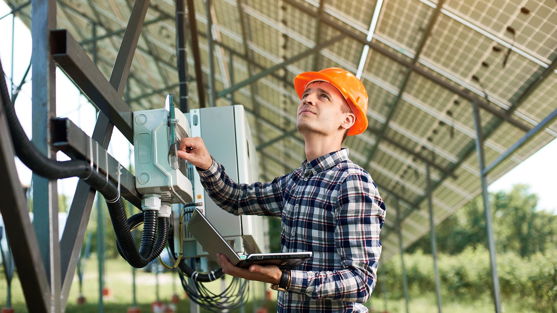 Male engineer in helmet and with laptop pushing button at the electrical equipment on solar power station, looking up. Science technology. Solar panels. Global warming. Green energy.