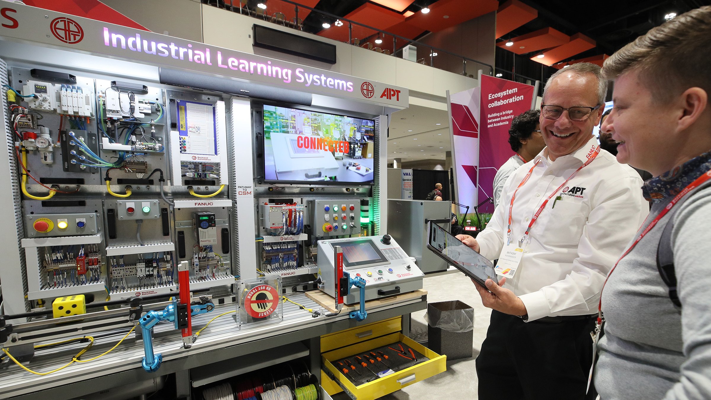 Two people looking at APT Industrial Learning Systems display. There are many products and they are interacting, the man is holding a tablet. 