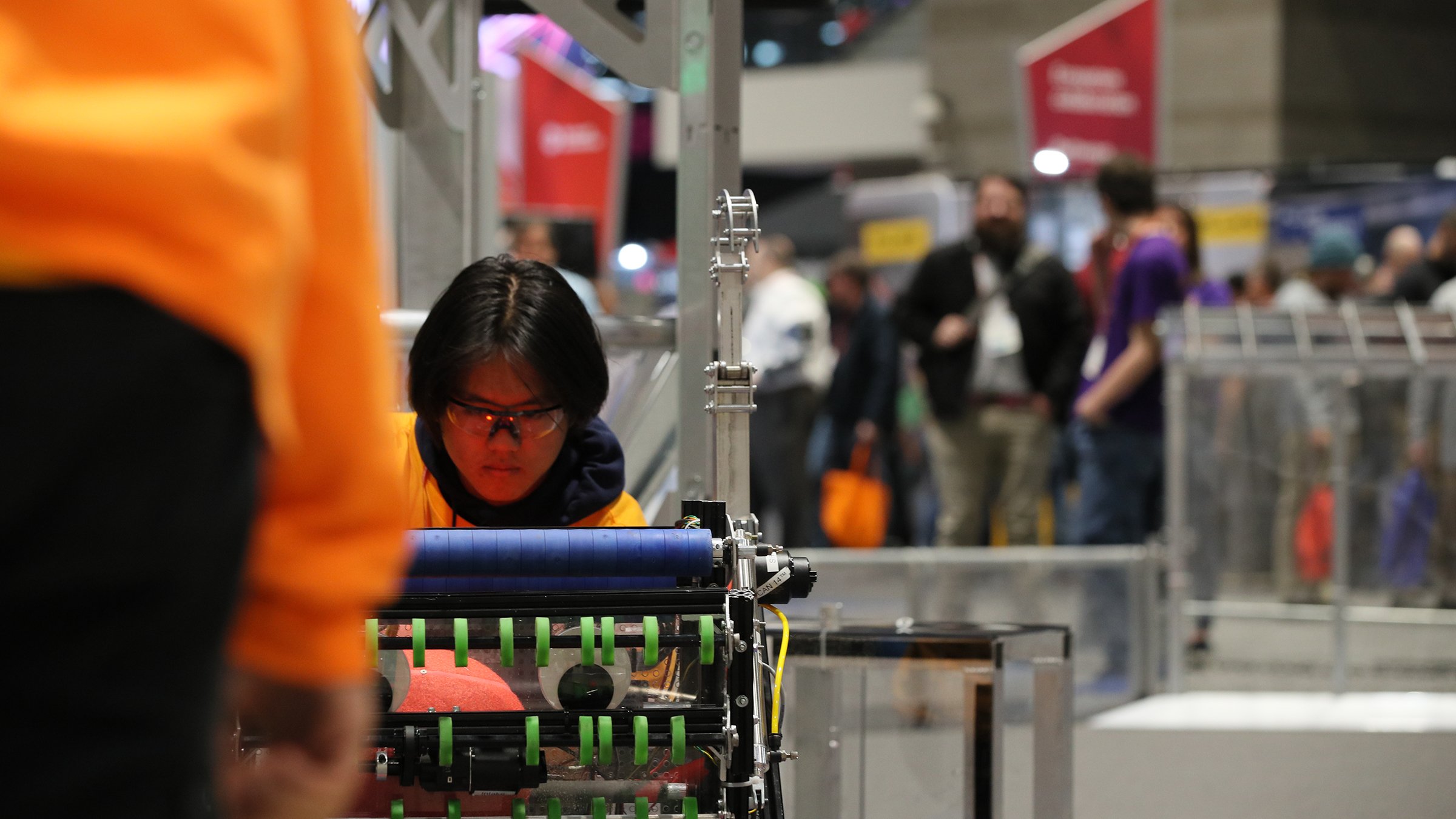 Person in safety glasses in foreground looking at machine. There are attendees on show floor in the background.