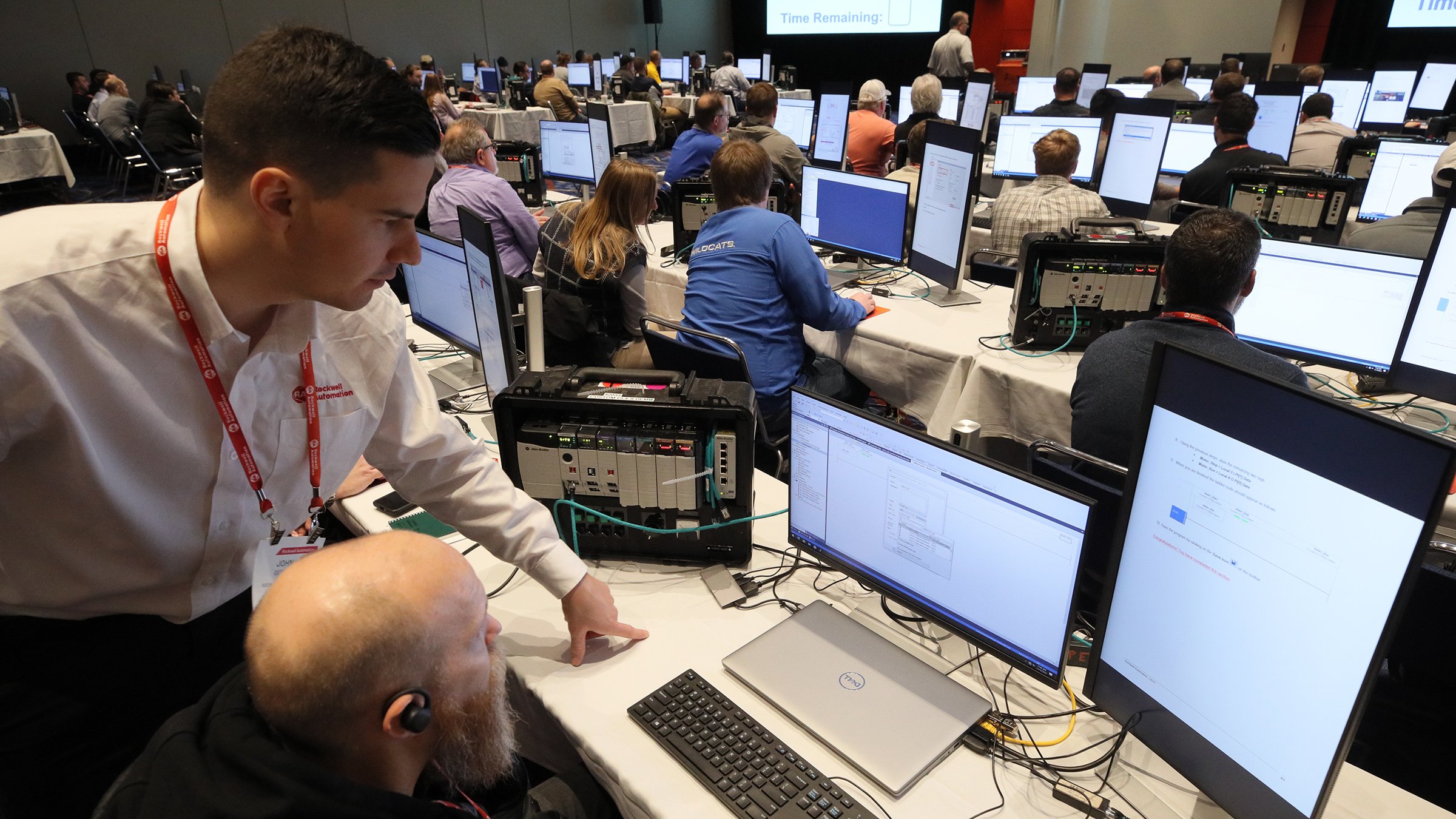Rockwell Automation instructor works with attendee in large session room with many workstations and people.