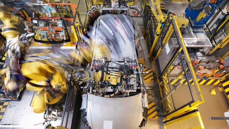 Braços robóticos movendo-se em alta velocidade enquanto trabalham na estrutura de um carro em uma fábrica automotiva.