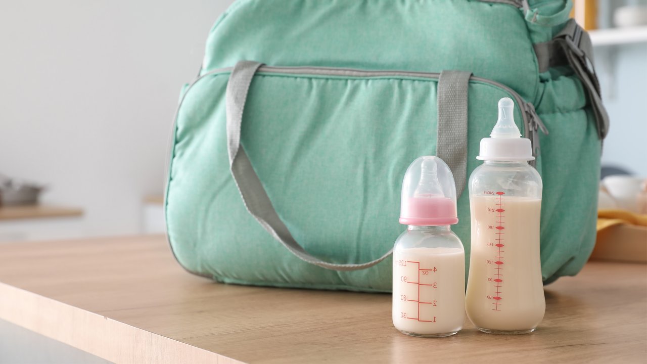 Two full baby bottles of milk in front of a teal diaper bag on a wooden table. 