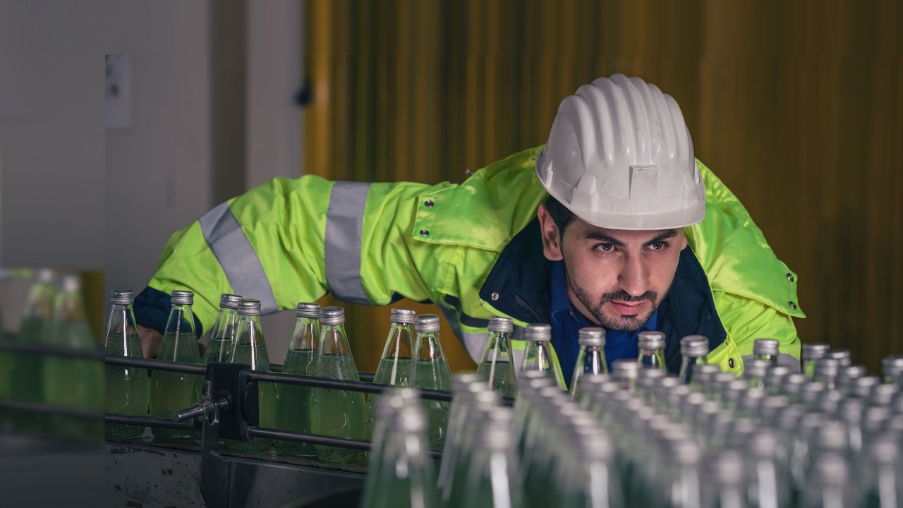 Worker in beverage production factory wearing hard hat leans over and checks the glass bottles on the conveyor