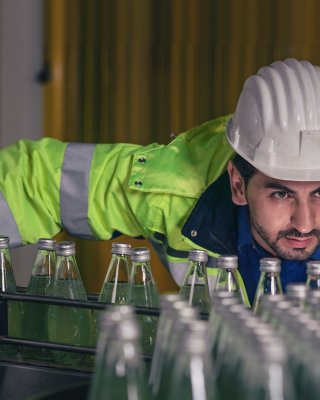 Worker in beverage production factory wearing hard hat leans over and checks the glass bottles on the conveyor
