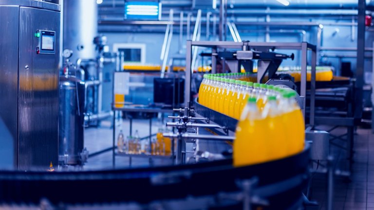Bottiglie con liquido arancione e tappi verdi che scorrono lungo un binario in un sistema di produzione con PC e monitor industriali.