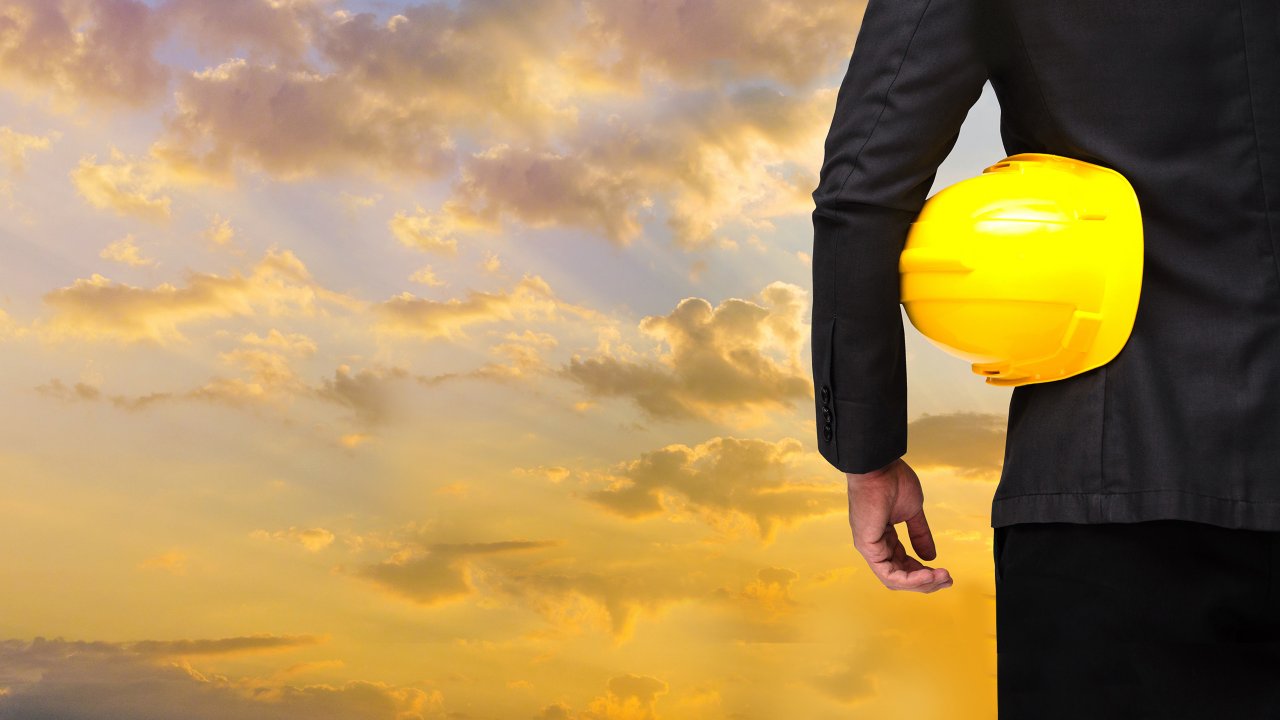 A person wearing a dark business suit holds a yellow hardhat under their arm while looking out at a sunset