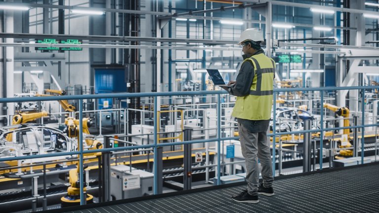 Male engineer in high visibility vest uses computer to check device cybersecurity while overlooking automotive plant