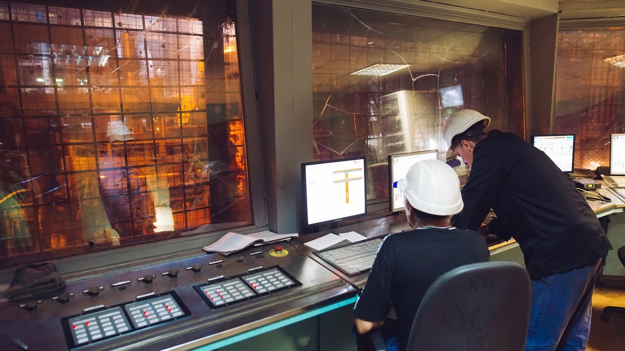 Two people wearing white hard hats looking at monitors in a control room above a steel mill
