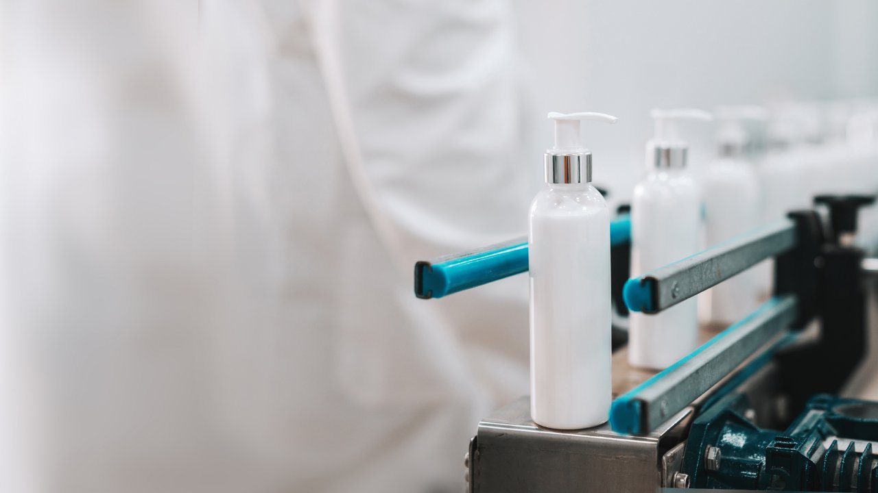  White cosmetic product bottles on manufacturing line with worker wearing a white lab coat in distance