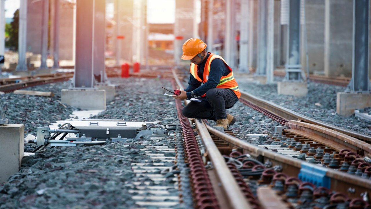 Man in hardhat with phone and clipboard kneeling near train track.