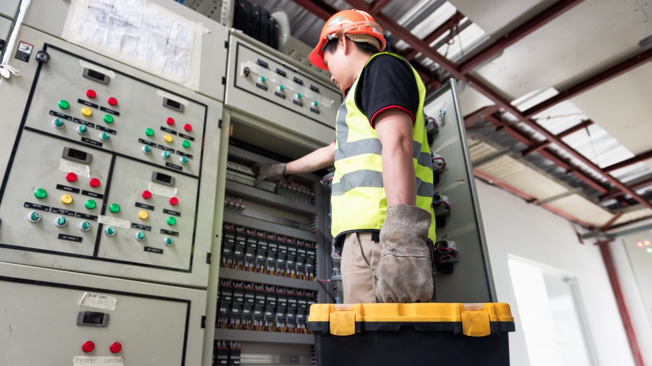 Electrician with toolbox working on fuse box in industrial control panel.